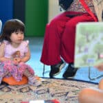 A girl sits on the floor, looking towards a book that someone is holding for her to see.